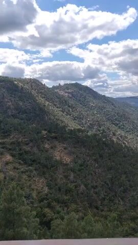 The video showcases a panoramic view of a mountainous landscape featuring a river winding through the valley. The foreground includes a portion of a railing, suggesting the viewpoint is from a deck or observation point. The hills are covered in dense green vegetation, with trees dominating the landscape. The river appears calm and reflects the sky above. The sky itself is dynamic, with patches of blue interspersed with white and gray clouds, creating a play of light and shadow across the scene. The overall impression is that of a serene and natural environment.