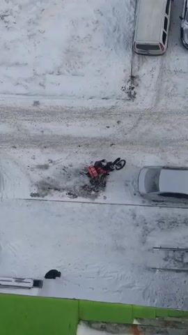 An aerial shot shows a motorcycle delivery person with a red thermal bag, traveling on a snowy, icy lot between parked cars. The surface is covered in snow and ice, with some cleared tire tracks. There are multiple cars parked in the lot, all covered in snow.  The delivery person appears to have some difficulty maintaining their balance as they navigate the slippery surface. The bottom edge of the video shows a green surface, likely part of a building.