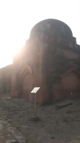 The video shows an ancient structure made of brown stone, seemingly a tomb or mausoleum, surrounded by a dirt and grassy area. Initially, the camera focuses on a part of the building with a dome-shaped roof and arched designs. As the video progresses, it pans out, revealing the entire structure and the surrounding environment, showcasing a pathway leading to the building, and two people walking around it. The sky is clear and bright, suggesting it's daytime. The architecture is old and looks worn, hinting at its historical significance.