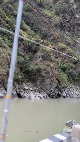 The video shows a river flowing alongside a steep, rocky mountainside covered with sparse vegetation. Power lines are strung across the mountainside. The water in the river is a murky, greenish-brown color. The sky is overcast. The camera appears to be mounted on a moving vehicle, as evidenced by the slight shaking of the image and the appearance of a barrier or railing in the foreground that seems to be moving along with the camera.