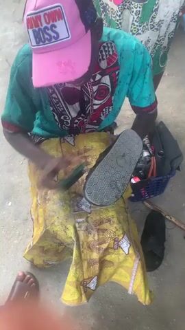 The scene depicts a man wearing a pink cap that says "I'M MY OWN BOSS," a turquoise shirt, and seated on what appears to be a yellow and brown cloth. He is engaged in shining a black sandal. He uses a brush and a green block to clean the sandal. A blue basket nearby contains tools and supplies. Two additional pairs of sandals and a hammer are on the ground around him. Other individuals are present in the background, but their actions are not focused on. The setting appears to be an outdoor area with a concrete surface.