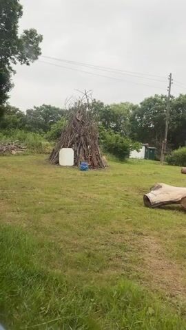 The video shows a grassy clearing with several objects. In the center of the frame is a large pile of branches, resembling a teepee or a large bonfire structure. To the left of the pile of branches is a white plastic barrel and a blue object, possibly a tarp or a bag. In the foreground, to the right, are two large, cut logs lying horizontally on the grass. Further in the background, beyond the pile of branches, are two small structures, one white and one green, with a utility pole and wires visible behind them. The area is surrounded by trees and foliage, suggesting an outdoor, possibly rural, setting. The sky is overcast. The video appears to be taken from a moving vehicle, as indicated by the slight blur and the perspective shifting slightly.