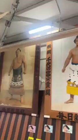 The video shows a series of framed portraits of sumo wrestlers. The portraits are displayed side-by-side along a wall, each one featuring a different wrestler in traditional attire. Japanese writing is present on the sides of the frames. The camera pans slowly across the portraits, revealing more wrestlers. Below the portraits is a vending machine. The overall lighting is bright.