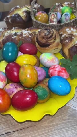 The video showcases a table laden with Easter treats. A yellow platter holds a collection of vibrantly colored Easter eggs in shades of red, blue, yellow, and various tie-dye patterns. A basket, lined with a lace doily, is filled with additional decorated eggs, a sprig of yellow flowers, and a small framed religious icon. Several freshly baked sweet breads, dusted with sugar and containing swirls of filling, surround the eggs and the basket. Green leaves are scattered around the arrangements, adding a touch of freshness to the display. The table surface is visible in the foreground, suggesting an indoor setting. The overall impression is one of festive celebration and traditional Easter preparations.