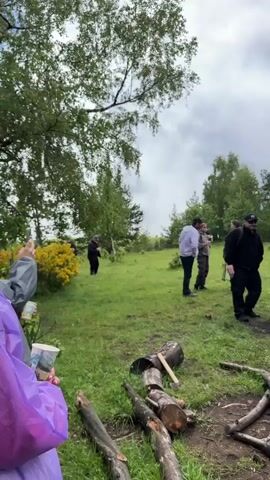 A group of people, appearing to be adults, are gathered in a grassy, wooded area. The sky is overcast. In the foreground, there are logs and branches, some of which appear to have been cut or broken. There is also evidence of a campfire, with burnt wood and rocks forming a circle. Several individuals are standing and talking amongst themselves. One person is holding a purple bag. Another person is in the background, standing near a bush with yellow flowers. The overall scene suggests an outdoor gathering or picnic.