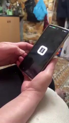 A person is holding a black smartphone with a gold-colored protective case. The screen shows an app with a woman's photo. The app has labels such as "Latest", "Created", and "Invite". The person's hands are visible, and they have fair skin. The background includes a floral patterned rug, a wooden cabinet, and other miscellaneous items.