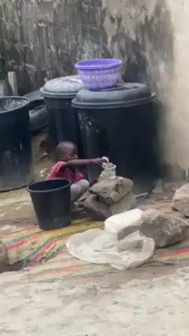 The video shows a small child squatting outdoors in what appears to be a courtyard or backyard area.  The child, dressed in a dark-colored shirt and light-colored pants, is focused on pouring liquid from a small white container into a glass placed atop a small stack of rocks.  Several large black plastic bins are positioned behind the child, and a black bucket sits to the child's left. A striped rug is placed under the child. The ground is concrete, with patches of dirt and debris. The background includes a weathered wall.