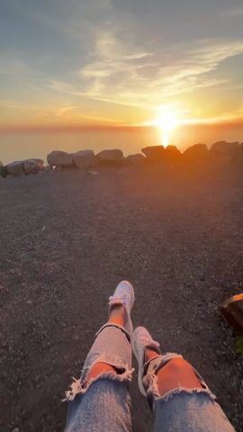 The video shows a person's legs and feet while sitting and facing a sunset over the ocean. The person is wearing white sneakers and ripped blue jeans. The ground is covered in gravel. There are large rocks along the edge of the ground. The sun is setting on the horizon over the water, and the sky is filled with orange and yellow hues.