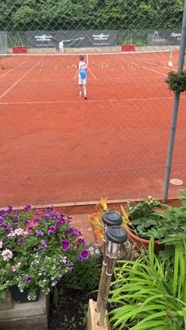 The video shows a person playing tennis on a clay court. The person is wearing white shorts and a white shirt with blue accents. The court is lined with tennis balls, and there are other people in the background, also on the court. The court is surrounded by a fence, and there are plants and flowers in the foreground.