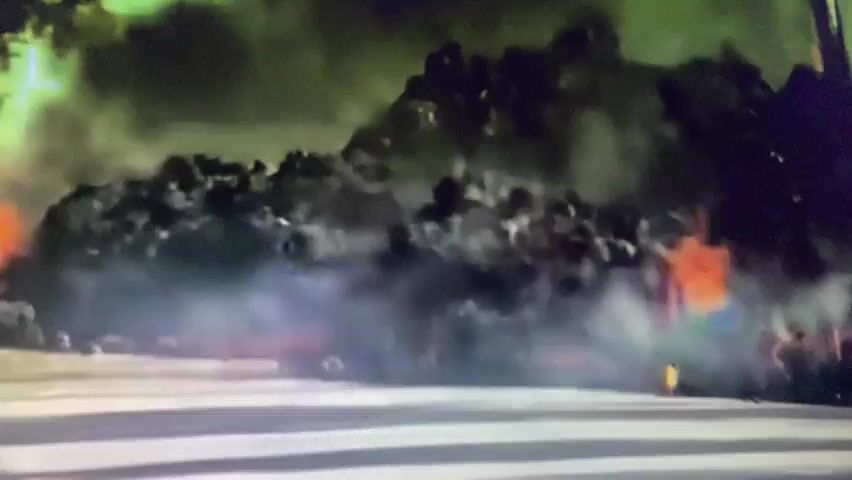 The video shows a pile of black material on fire, with flames and smoke visible. The material appears to be piled up and is burning from within. There is a greenish-yellow haze above the fire, possibly smoke or other atmospheric conditions. The foreground is a blurry gray surface, possibly a road or pavement. The overall scene suggests a significant fire.
