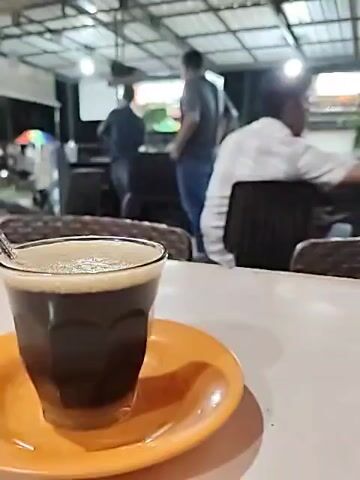 Here is a description of the video:
The video shows a glass of dark coffee on an orange saucer placed on a white table. There is a small spoon inside the coffee glass.  In the background are several people in a semi-outdoor area, possibly a cafe or restaurant.  They are standing or sitting.  The lighting appears to be from overhead fluorescent lights.