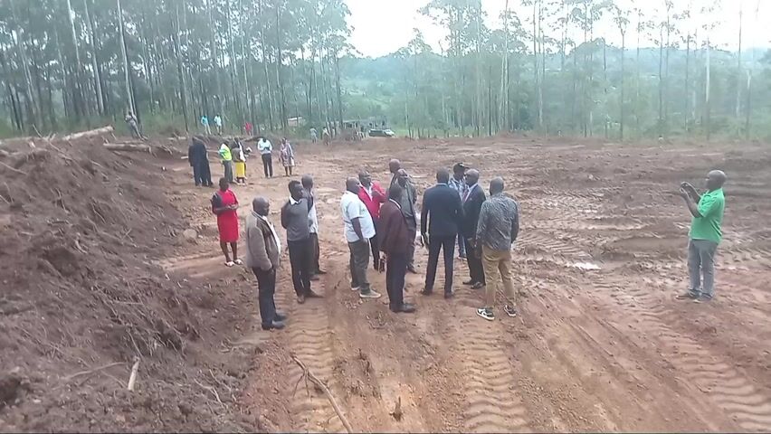A group of about 17 people are standing in a muddy clearing near a forest. Most of the individuals are men, but there are also several women. The men are wearing various outfits, including suits, dress shirts, casual shirts, and pants. The women are wearing dresses. One man on the right is taking pictures with his phone. The group seems to be engaged in a conversation or discussion. The area is a clearing of red dirt with deep tire tracks. In the background, there is a dense forest of tall, thin trees. The sky is overcast, and the lighting is subdued. The location appears to be rural and possibly an area that has been cleared for development or construction.