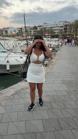 The video shows a woman with long dark hair and a white dress standing on a paved walkway next to a body of water. Several boats are docked in the water, and buildings and palm trees are visible in the background. The woman is looking at the camera and touching her hair. In one shot, she is holding a black purse. The sky is cloudy.