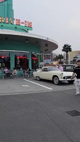 The video shows a group of people at a retro-themed drive-in restaurant. The setting appears to be a sunny day, likely late afternoon or early evening, given the soft light. The restaurant itself is a prominent feature, with a curved teal facade and a large sign reading "DRIVE-IN" in neon letters. Below the sign, the word "Hamburgers" is also visible in neon.

Several individuals are present. In the foreground, a young couple walks past the camera. The man is wearing a black t-shirt with white lettering and light-colored pants, while the woman wears a plaid skirt and a light-colored top. They appear to be casually strolling and conversing.

In the middle ground, a classic cream-colored convertible, possibly a 1950s-era car, is parked in front of the drive-in. A young woman in a pink outfit is standing on the hood of the car, posing and looking towards the camera. Another person, partially visible, is taking a picture of her.

Inside the drive-in, through the large windows, several other patrons can be seen sitting at tables. Some appear to be young women, enjoying their time at the establishment. The overall atmosphere seems relaxed and cheerful, with a nostalgic vibe due to the retro styling of the restaurant and the car. The presence of palm trees in the background hints at a warm climate, and the location context suggests this is in Tongzhou District, China.