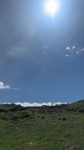 The video shows a low-angle view of a grassy, rocky hillside against a bright blue sky with scattered white clouds. The sun is high and shining intensely. Two figures are visible on the crest of the hill, silhouetted against the sky. The camera appears to move slightly, adjusting the framing of the scene. The overall environment suggests an outdoor, sunny day in a natural setting.