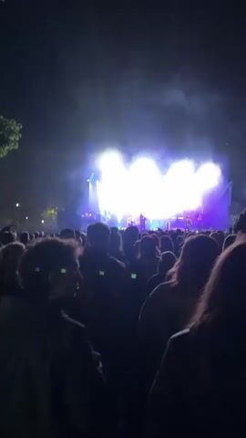 A live music concert is in progress. A band is performing on stage, bathed in bright white and purple lights. The crowd in front of the stage is dense, with many people holding up their phones to record the performance. A screen behind the band displays a large image of a man with long dreadlocks. The setting appears to be an outdoor venue at night, with some trees visible on the left side of the frame.