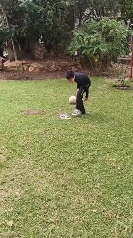 A young boy, dressed in a black long-sleeved shirt and black pants, is playing soccer on a lush green lawn. He is wearing white sneakers with blue accents. A light-colored soccer ball is in front of him. The boy appears to be in mid-action, either about to kick the ball or having just kicked it, with one leg raised.

The setting is an outdoor grassy area, possibly a backyard or a park, with trees and some rocky landscaping in the background. Behind the lawn, there is a building with a wooden door and windows, suggesting it's a residential or recreational area. The lighting suggests it is daytime. The overall mood seems casual and active, with the boy engaged in a sporting activity.