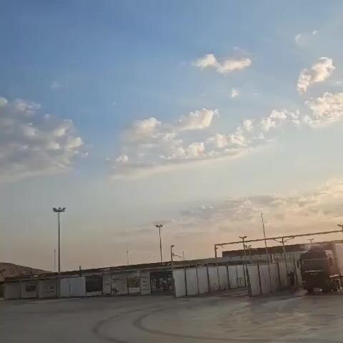 The video shows an outdoor view of a paved area with some structures, likely industrial or storage units, along the perimeter. In the foreground, the concrete surface appears wet, reflecting the light. A dark-colored truck is parked in the middle ground to the right. The sky is visible with scattered white clouds and the sun is setting, casting a warm glow and visible rays across the scene. The time stamp indicates the video progresses from 00:00 to 00:05.