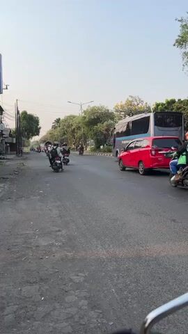 This video shows a street scene with several people riding motorcycles. The camera appears to be mounted on the back of a motorcycle, giving a view of the road ahead. In the foreground, the back of a motorcycle and the rider's legs are visible. The road is paved and appears to be in moderate condition. Several other motorcycles are on the road, some with single riders and others with two people. In the background, there are trees, buildings, and a large bus. The sky is clear and appears to be daytime. The overall impression is of a typical street scene in a populated area with a lot of motorcycle traffic.