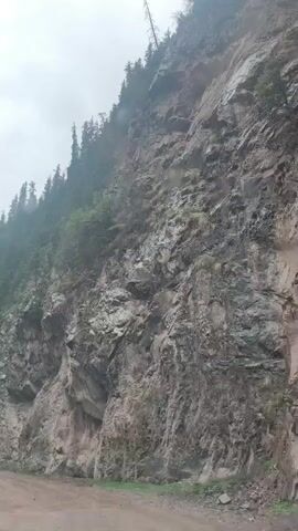 The video, filmed from inside a moving vehicle, shows a rugged, mountainous landscape in Dzhetyoguz, Kyrgyzstan. The main subject is a steep, rocky cliff face covered in patches of dark green vegetation and conifer trees, stretching upwards towards an overcast, grey sky. The rock face appears wet, suggesting recent rain or ongoing light precipitation, which is corroborated by the blurred motion of windshield wipers in the foreground. The vehicle is traveling on a dirt road that runs parallel to the base of the mountain. In the distance, another vehicle can be vaguely seen on the road. The scene depicts a journey through a wild, natural environment on a cloudy day.