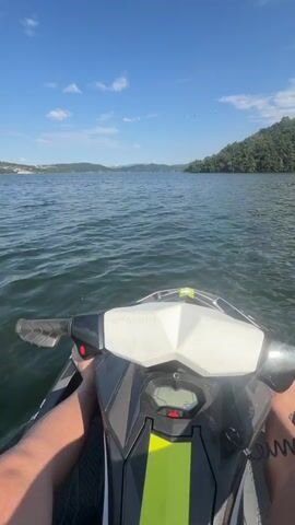 The video shows a first-person perspective of someone riding a jet ski on a large body of water. The rider's legs are visible in the foreground, with their left hand on the handlebars. The jet ski is moving, creating a wake. In the background, there are rolling hills covered in trees, and a distant shoreline with some buildings. The sky is blue with scattered white clouds. The video is shot on a bright, sunny day. The jet ski's digital display shows the speed as 28 km/h.