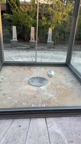 The video shows a glass-enclosed structure with a sundial on the floor. A metallic sphere is suspended from the ceiling by a wire and swings back and forth like a pendulum. The sundial has radial lines emanating from a central point and circular markers around its perimeter. Outside the structure, there are trees, buildings, and three tall, white, conical sculptures. The ground outside is paved with stone. The lighting suggests it is daytime, with sunlight casting shadows.