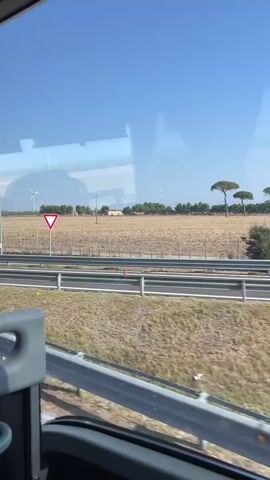 The video is taken from the perspective of a passenger inside a vehicle, likely a bus, looking out of the right-side window. The view captures a roadside landscape under a clear blue sky. In the foreground, a portion of the vehicle's interior is visible, including a grey seat with a red handle and a portion of the window frame. Adjacent to the window, the grey seatback of another seat is visible. The window itself reflects parts of the interior of the vehicle. Outside the window, a highway or major road is present, with a metal guardrail separating it from a dry, brown field. White lane markers are visible on the road. In the distance, several wind turbines are scattered across the field. A tall, thin streetlamp stands near the road. A triangular yield sign with a red border and a white interior is posted on a pole in the field. The field is enclosed by a wire fence with wooden posts. Some sparse vegetation, including a bush or small tree, is visible in the field. A dark-colored car is partially visible on the road in the middle distance, appearing to be driving in the same direction as the viewer's vehicle. The overall impression is a scenic drive through a rural area on a sunny day.