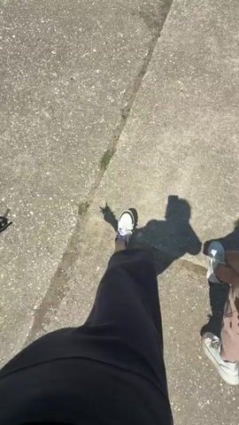 The video shows a group of people walking on a sidewalk. The camera is focused on the ground and shows the shoes and shadows of the people walking. The sidewalk is made of concrete and there is some debris on the ground. The sun is shining, casting long shadows.
