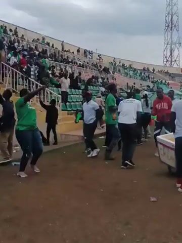 The video shows a group of people dancing in front of stadium seating. The seats are filled with fans and many are wearing green. Some of the dancers are wearing shirts that say "NYAKWAR NYADEDE." One person wears an Indiana sweatshirt, and another pushes a white cart with "CREAM" on the front. A tall antenna is visible in the background.