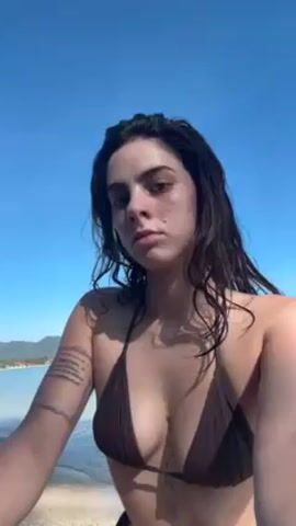 A young woman walks toward the camera on a tropical beach. She has dark, wet hair and is wearing a brown string bikini. She has tattoos on her left arm and torso. The beach appears sandy, with clear blue water and a distant, lush coastline. The sky is also clear and blue. She stops briefly and then continues walking into the water.