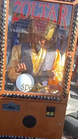 The video shows a Zoltar machine in an outdoor setting, possibly a market. Zoltar is a fortune-telling machine with an animatronic figure inside, dressed as a mystic. The machine is brown and has a glass front displaying the Zoltar figure. The figure is wearing a gold turban, yellow shirt, grey patterned vest, and a large silver chain. It has a beard and appears to be holding its hands over a white, opaque crystal ball. The word "speak" is written on the glass near the crystal ball. Reflections on the glass show the outside environment, including other vendors and people walking by. The camera moves slightly, zooming in on the Zoltar figure.