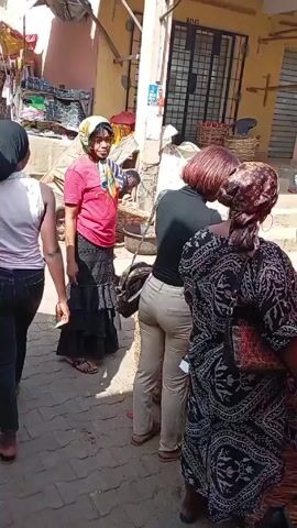The video shows an outdoor market in daylight, with several people shopping at produce stands. Several women are visible, browsing tomatoes and peppers. One woman in a black and white patterned dress and headscarf stands at a stand. Another woman in a red shirt and long, black skirt is standing near the stand. A third woman, wearing khaki pants and a black shirt, is also looking at the produce. Several baskets of produce are displayed. The vendors are interacting with the customers, possibly discussing prices or quantities. The environment appears to be a bustling market area, with other people and objects in the background.