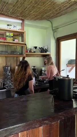 Two women are preparing drinks behind a coffee bar counter. The woman on the left has long wavy brown hair, is wearing a black top and a dark apron. The woman on the right has long blonde hair and is wearing a pink top and dark shorts. They are working side-by-side behind a stainless steel counter. An espresso machine, a grinder, and other small appliances are arranged on the counter. Behind them are shelves with boxes, cups, and bottles. A wooden counter separates the workers from the rest of the room. The walls are a light green color. The ceiling has a woven bamboo-like material.