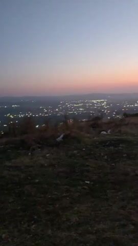 A very low-light, handheld video shows a dimly lit city skyline at dusk from a hilltop. The sky transitions from a deep blue at the top to shades of pink and orange near the horizon. The city lights appear as small, blurred yellow dots spread across the landscape. The foreground is dark and grassy, with some scattered debris that is difficult to discern. There are a few dark, indistinct shapes of trees and possibly bushes on the hilltop. The camera is mostly stationary but does exhibit minor shaking.