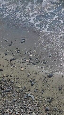 An aerial view of a shoreline shows waves crashing on a pebble beach. The water is clear and reflects sunlight. The beach is covered in small stones and sand.