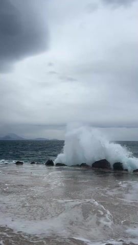 The video shows an outdoor scene of an ocean on a cloudy, overcast day. The sea is dark blue and choppy, with white waves crashing against a group of dark rocks near the shoreline. The waves create large splashes of white water. The foreground is a sandy area with foamy water lapping over it. The sky is filled with gray clouds, and distant land is visible on the horizon. The video is static, with no camera movement.