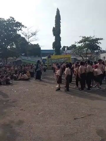 In an eye-level wide shot captured outdoors during daylight, a large group of children and a few adults are gathered in what appears to be a schoolyard or open space. The children are mostly clad in uniforms consisting of brown tops and dark shorts or skirts, and are divided into two groups. One group is seated on the ground in a semi-circle, while the other is standing in rows.

Several adults, predominantly women wearing headscarves and long dresses, are interspersed among the children, seemingly supervising or instructing them. One woman is standing near the seated group, possibly leading a discussion or activity. The ground is uneven and covered with loose dirt and gravel. There are some white markings on the ground, possibly indicating boundaries or markers for activities.

In the background, there is a mix of trees, some still retaining their leaves and others appearing bare. A tall, slender tree stands prominently on the right side of the frame. A light blue truck with some yellow accents is also visible in the background, parked behind the group. Further back, there are some buildings and a sign with the word "APEL" visible.

The overall atmosphere appears to be of a school event or gathering. The lighting is bright, with the sun casting shadows on the ground.