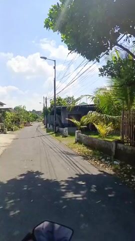 A first-person view from a vehicle shows a narrow, paved road in a residential area. On the right, numerous overhead utility lines run along with several utility poles. Houses with various fences and some foliage are visible on both sides of the road. The sky is bright blue with scattered white clouds. The sun causes a significant lens flare in the upper right quadrant of the screen. A small pile of dirt is visible on the left side of the road. A person on a scooter is approaching in the distance. The vehicle travels straight down the road.