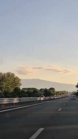A view from a moving vehicle on a highway. Cars are visible in both lanes, traveling in the same direction as the camera. A metal guardrail runs along the left side of the road. Trees and vegetation line the area beyond the guardrail, with a mountain range in the distance under a pale blue sky with scattered clouds. The sun appears to be low in the sky, casting long shadows and creating a soft, warm light.