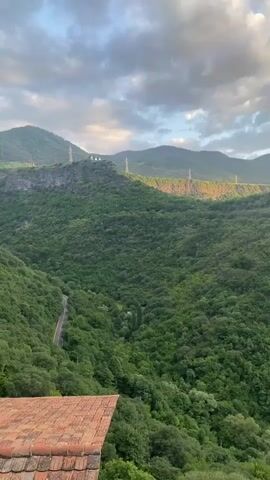 This is a video of a mountainous, forested landscape, likely taken in Haghpat, Armenia, during the late afternoon or early evening. The sun is casting long shadows and illuminating the tops of the trees and a distant ridge with a warm glow. The sky is partly cloudy, with dramatic, textured clouds that are catching the sunlight.

The scene is dominated by rolling, verdant hills covered in dense green forest. A winding road is visible in the valley below, disappearing into the trees. In the distance, a series of tall, slender pylons or power transmission towers are spaced along the ridge line, hinting at the presence of infrastructure within this natural setting. Further along the ridge, a historic stone structure, possibly a monastery or a fortified building, stands prominently on the crest of a hill, silhouetted against the sky.

In the foreground, the corner of a terracotta-tiled roof is visible, suggesting the vantage point is from a building or structure overlooking the valley. The overall impression is one of natural beauty and historical significance, with the dramatic lighting and vast landscape evoking a sense of awe and tranquility. There is no visible text in the video.