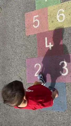 The video shows a young child playing hopscotch on an asphalt surface. The hopscotch grid is painted with colorful squares numbered from 1 to 10. The child, wearing a red shirt and sandals, is standing on square number 4. The sun casts a shadow of the child onto the hopscotch grid, with the shadow also appearing on square number 6. The child appears to be in the process of playing the game, possibly preparing to jump to the next square. The environment is outdoors, with the asphalt ground and the brightly colored hopscotch grid being the primary visual elements.