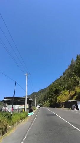 The video shows a view from a vehicle moving along a paved road on a bright, sunny day with a clear blue sky. The road curves slightly to the right and is bordered by a grassy shoulder on the left and a hillside covered with trees on the right. A utility pole with multiple wires runs along the left side of the road. On the left shoulder, there are some structures resembling small roadside stalls or buildings, with some bags and possibly produce visible. A white car is parked or moving slowly ahead on the right side of the road. The overall environment appears to be a rural or mountainous area.