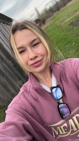 A young woman is recording a selfie video outdoors. She has fair skin with freckles, blonde hair pulled up in a casual bun, and is wearing a pink sweatshirt with a white t-shirt underneath. Sunglasses are hanging from the neck of her sweatshirt. The background shows a field, part of which is green grass and part is dark soil, possibly mud. The sky is overcast. The woman smiles at the camera and raises one hand in a wave.