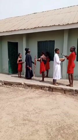 The video shows a group of people enacting a scene outdoors in front of a light green building. There are six people visible. Four of them appear to be children, three of whom are wearing red clothing, and one is dressed in white. Each is holding a wooden tray. A person dressed in a long black robe and a black hat stands next to the children in red, holding a decorated staff. A woman in a red dress is holding a microphone and appears to be interviewing the child in white. The building behind them has a light-colored roof and a dark green door on the left. The ground is dirt or sand.