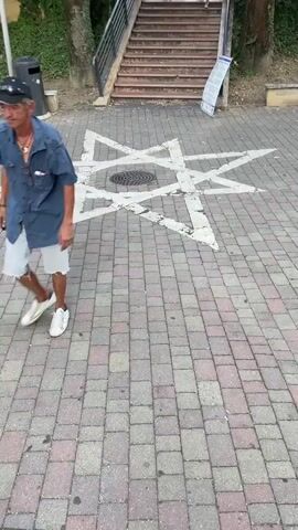 The video shows a street scene with a large Star of David painted on the ground. The star is white and appears to be made of paint or a similar material, and it is painted over a cobblestone pavement. There is a manhole cover in the center of the star. To the left of the star, there is a black trash can and a person walking in white sneakers. In the background, there are stairs leading up a hill, with trees and some railing along the stairs. The video is taken from a low angle, looking down at the ground. The lighting suggests it is daytime.