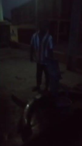 The scene is a dimly lit outdoor area where a person is standing with a long blade, possibly a machete or knife. The person is wearing a vertically striped shirt and dark pants. They appear to be attending to a large, dark object on the ground. The object seems to have a curved shape, possibly a pipe or large piece of metal. There are other objects and shadows in the background, but they are hard to make out due to the low light. The ground appears to be wet or damp.