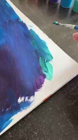 The video shows a close-up shot of a hand holding a paintbrush, applying bright green paint to a canvas. The canvas is partially covered with strokes of blue and purple paint, suggesting an abstract artwork. The background is out of focus but appears to be a countertop with some blurry objects, possibly paint bottles, on it. The lighting is bright, indicating it could be daytime. The scene is set in a home or studio environment where someone is engaged in painting. The focus is entirely on the artistic process.