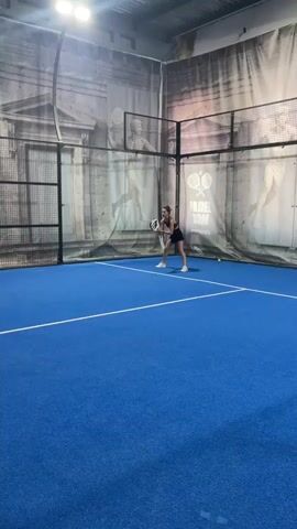 A woman in athletic attire, wearing a dark tank top and shorts, stands on a blue padel court, holding a padel racket. She is in a ready stance, with her knees bent and the racket held in front of her. The court is enclosed by glass walls and metal fencing, with a net dividing it in half. The background features a large mural depicting classical architecture and figures. Another woman, wearing a light-colored dress, stands on the opposite side of the net, also holding a racket. A few yellow padel balls are visible on the court. The lighting is bright, suggesting an indoor facility. The video captures a moment of anticipation before or during a padel game.