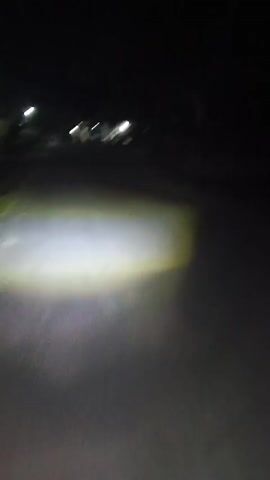 The video shows a first-person perspective of someone riding a motorcycle at night on a paved road. The motorcycle's headlight illuminates the road ahead, casting a bright beam of light. The surrounding environment is dark, with some trees and buildings visible in the distance. There are also streetlights along the road. The motorcycle appears to be moving at a moderate speed, as evidenced by the motion blur in the video. The scene is relatively still, with no other vehicles or pedestrians visible on the road.