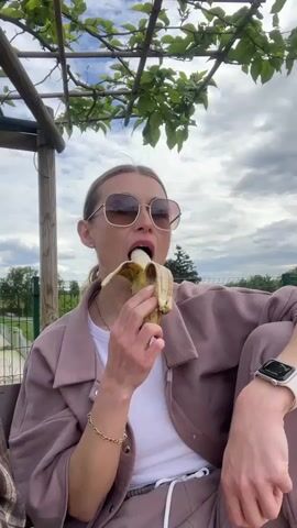 A woman sits under a wooden pergola with leafy vines overhead, eating a banana. She is wearing a mauve-colored tracksuit, a white t-shirt, and sunglasses. She has a gold bracelet and a smartwatch on her left wrist. The background features a park with green grass, a fence, and trees under a cloudy sky. Voices of children can be heard in the background.